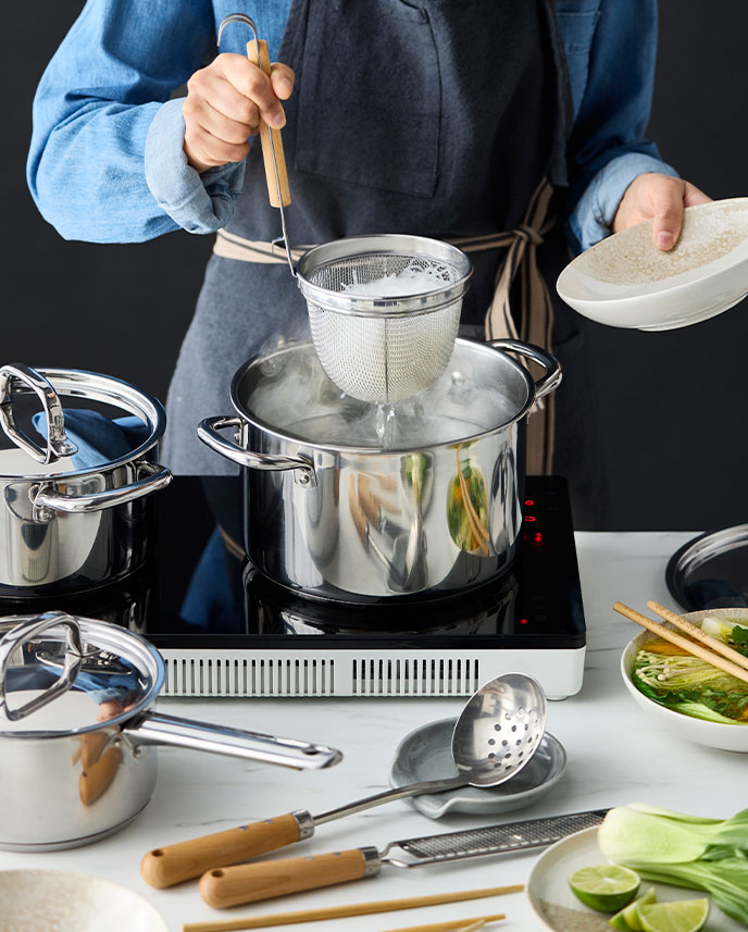  A person cooking on a white TILLREDA portable induction hob with two cooking zones while using a VARDAGEN noodle strainer.