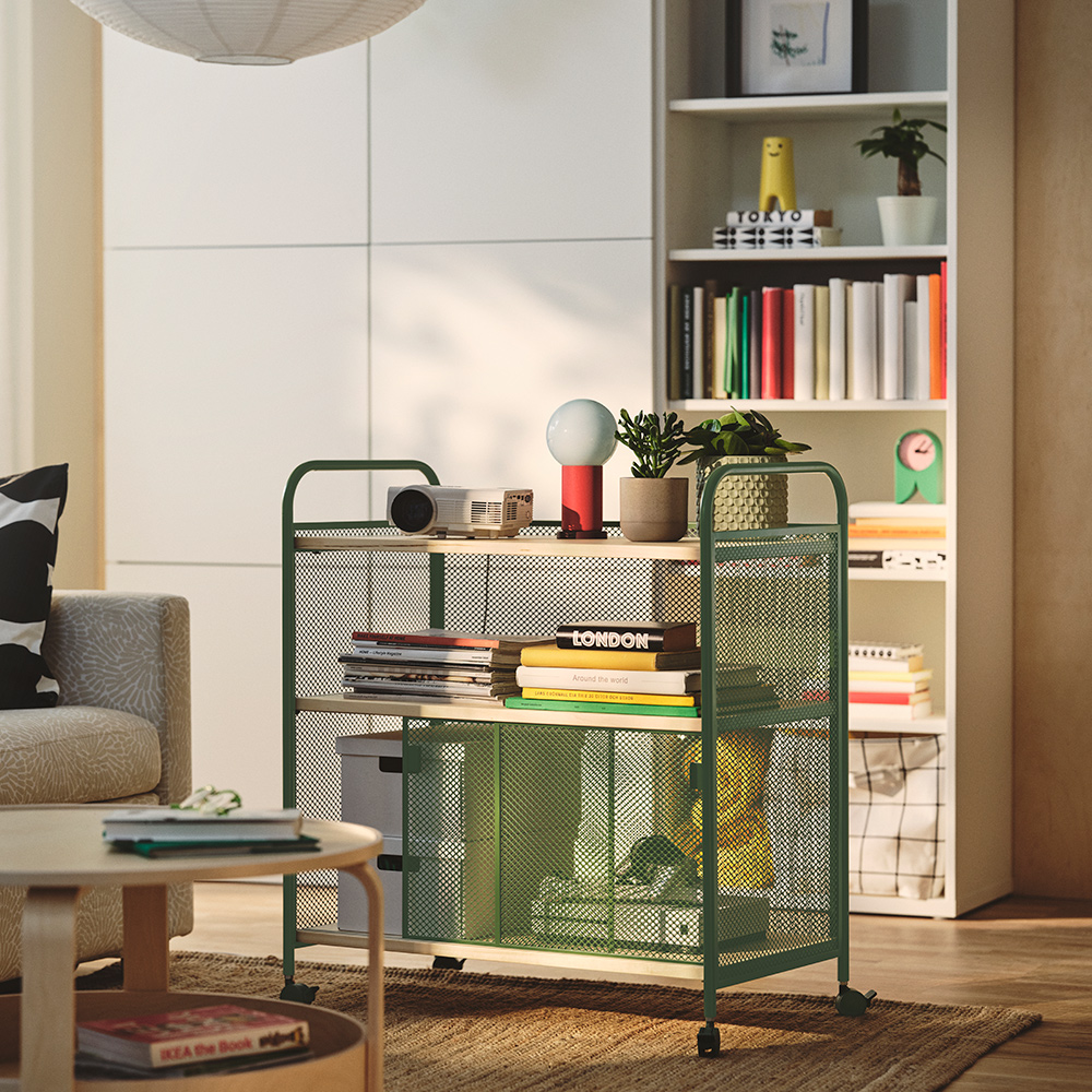 Aliving room with a dark green NALMAKARE cabinet on castors holding boxes and plants. A bookcase in the background.