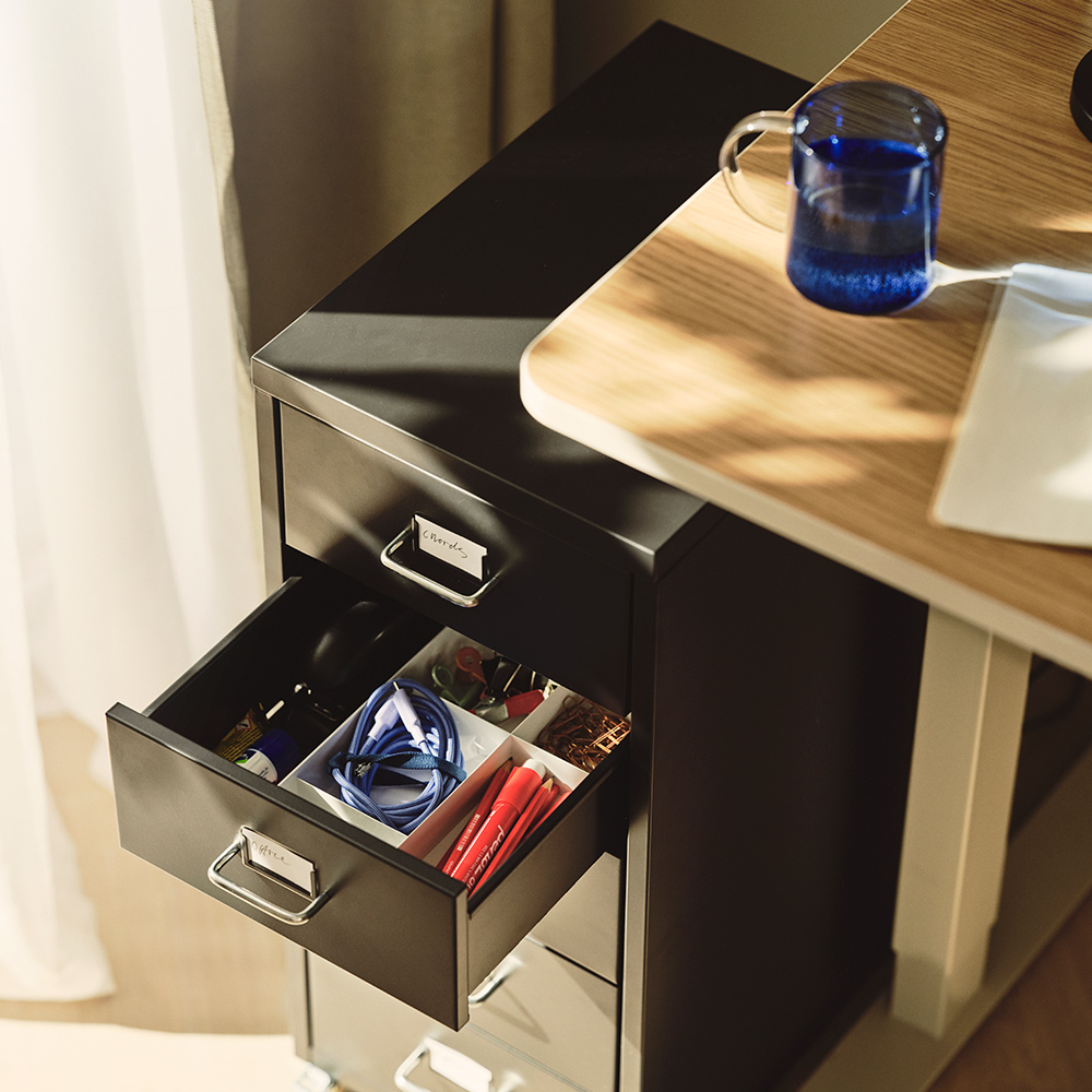 A close-up of a black HELMER drawer unit on castors with one drawer open, showing neatly arranged items inside.
