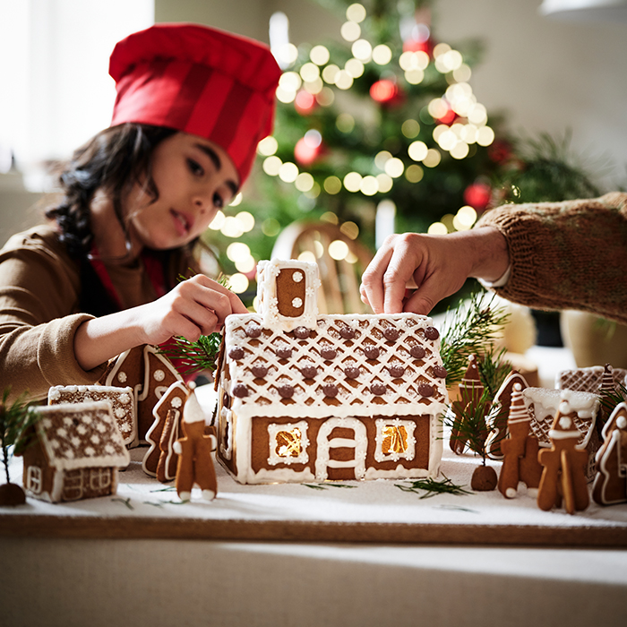 Casa de jengibre VINTERSAGA decorada con glaseado y rodeada de figuras y pequeñas casitas de galleta, mientras se termina de decorar sobre una mesa de temporada festiva.