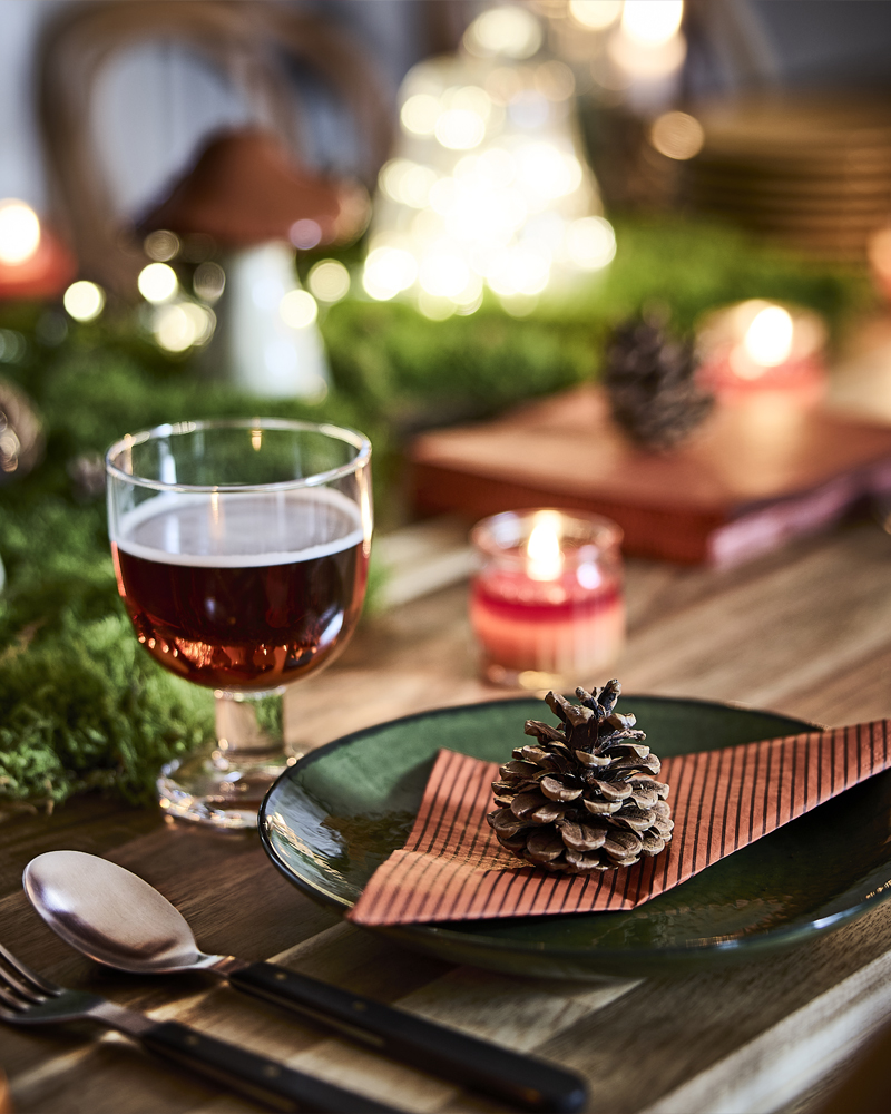 An autumn-themed table setting with a pinecone resting on a folded HÖSTAGILLE paper napkin placed on a green glass plate.