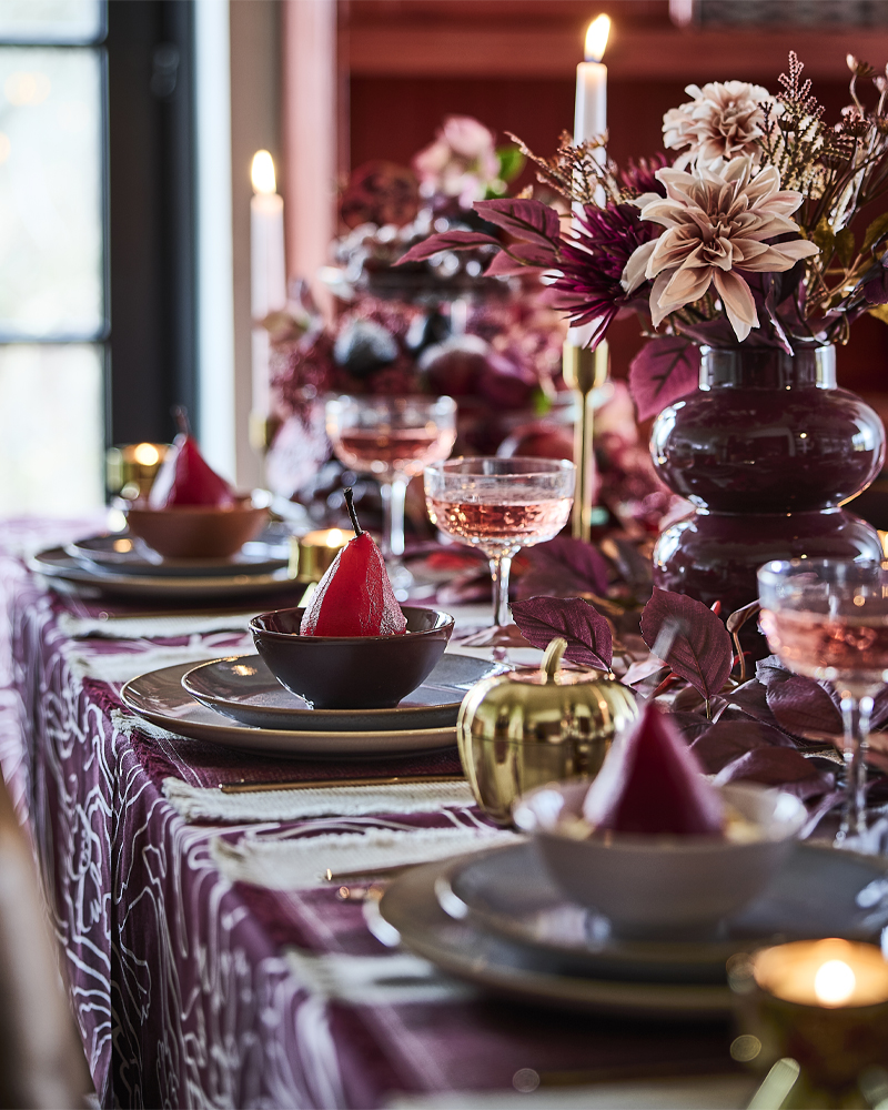 A pink and red table setting with lit candles, HÖSTAGILLE artificial bouquets in vases and poached pears in bowls.