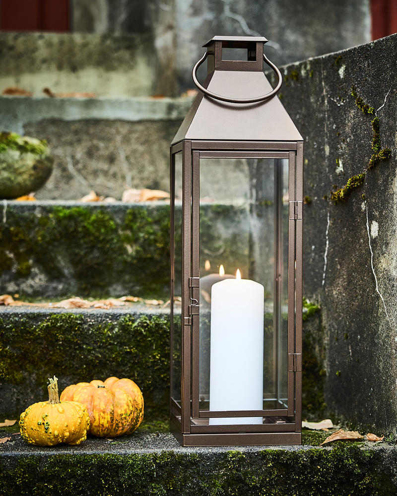 A lit white pillar candle inside a HÖSTAGILLE lantern next to two mini pumpkins on an outdoor staircase.
