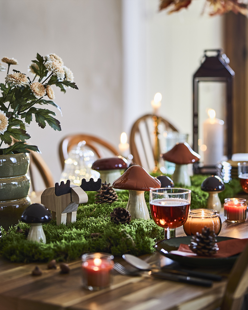 A woodland-themed dining table is set with HÖSTAGILLE decorative mushrooms and moose, lit candles, moss and pinecones.