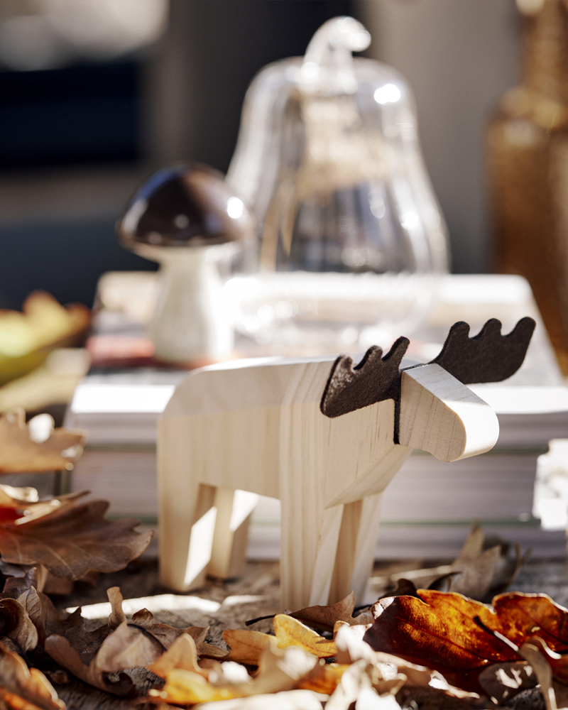 A HÖSTAGILLE decoration moose stands amidst dried leaves, next to a pile of books with a HÖSTAGILLE decorations on it.