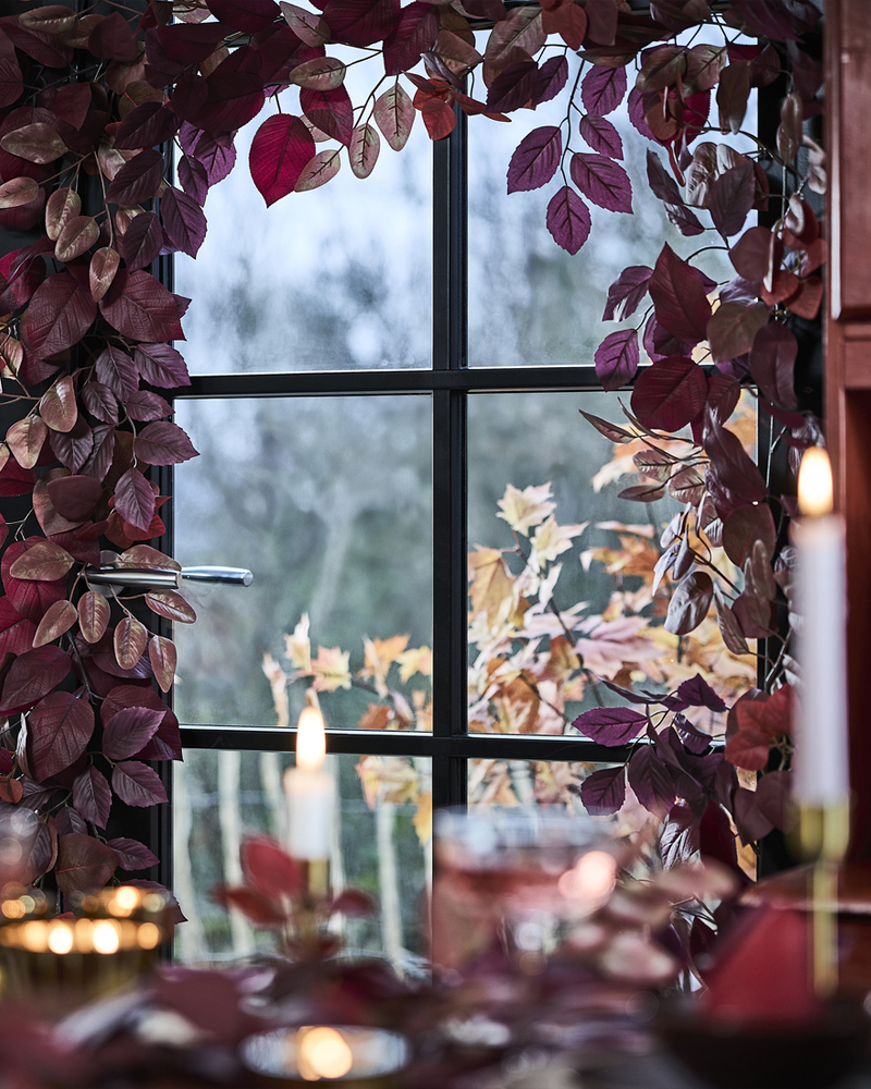 A glass window is richly decorated with red SMYCKA artificial garlands, next to a dining table with lit candles.