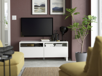 Mueble de TV blanco con televisor, lámpara y libros contra una pared malva, flanqueado por una planta y sillones amarillos.