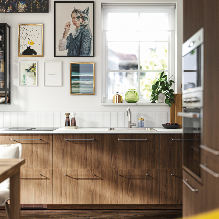 Cocina con muebles de madera, encimera blanca, ventana iluminada y galería de cuadros.