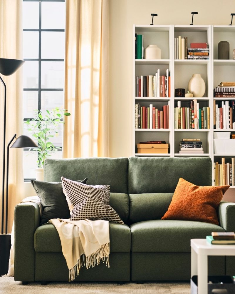 A light living room with a grey-green MALMKÖPING two seat sofa flanked by a bookcase, a floor lamp and plants