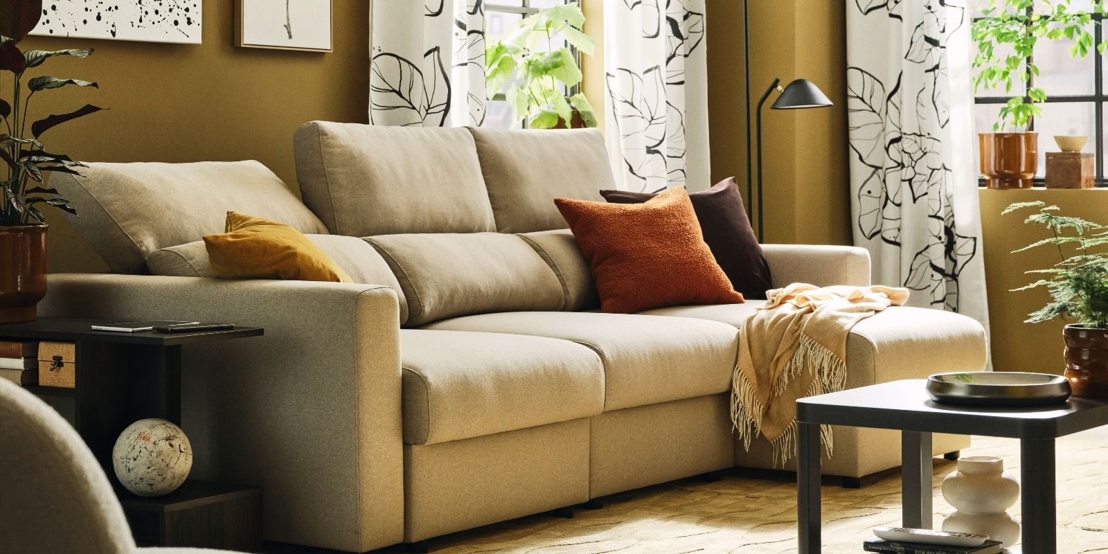 A light living room with a light-beige MALMKÖPING three seat sofa with chaise longue flanked by side tables and plants.