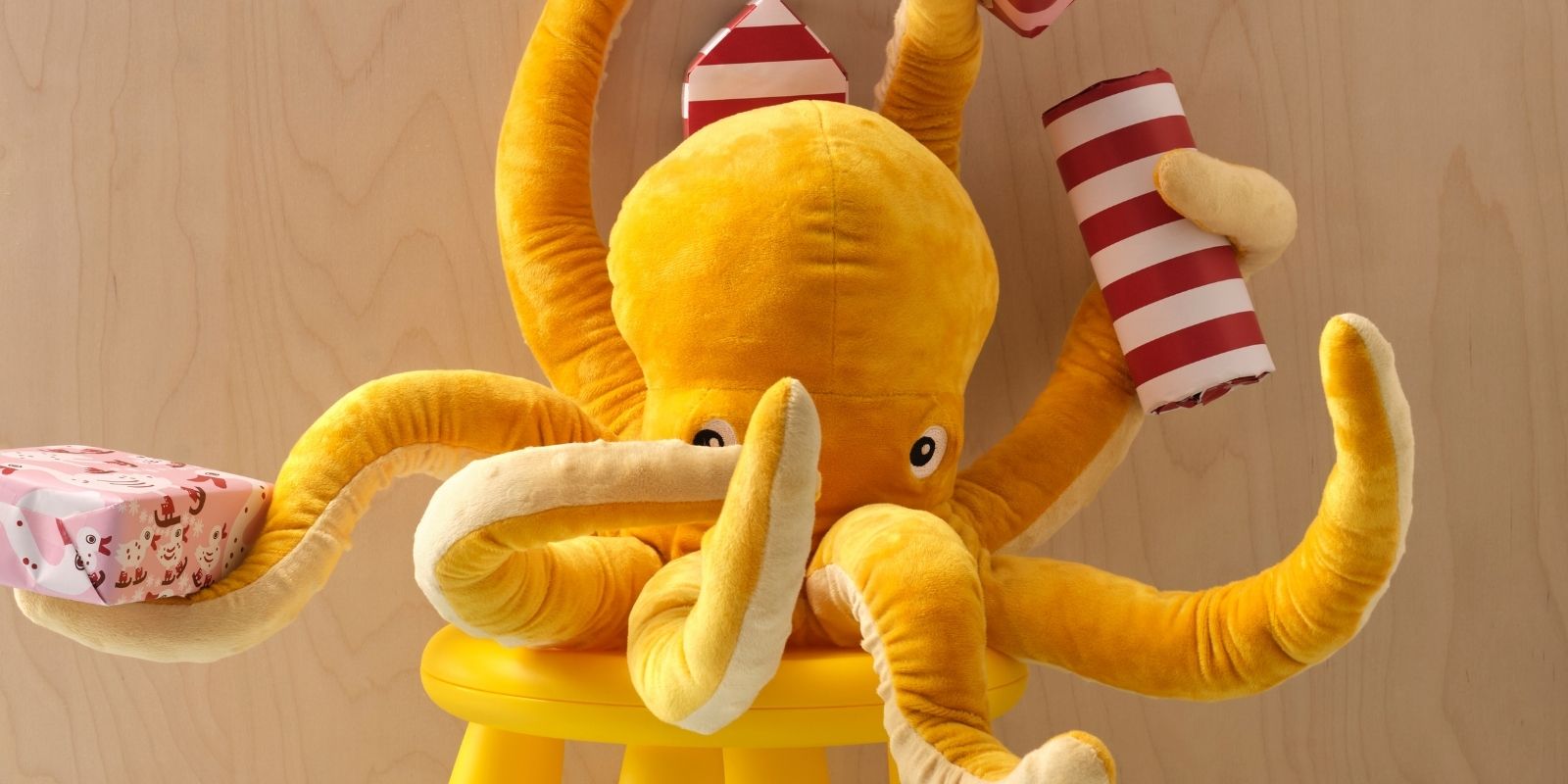 A yellow BLÅVINGAD soft toy octopus is sitting on a MAMMUT stool, holding several wrapped gifts in five of its eight arms.