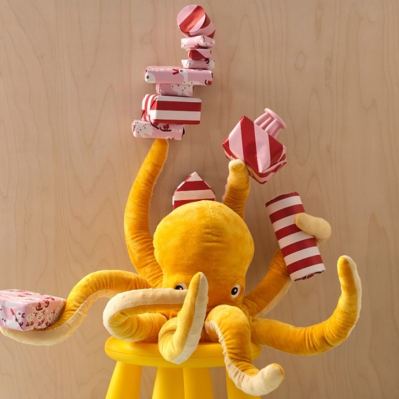 A yellow BLÅVINGAD soft toy octopus is sitting on a MAMMUT stool, holding several wrapped gifts in five of its eight arms.