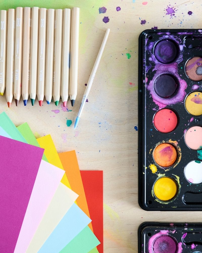 Coloured paper, a set of MÅLA pencils and watercolour box, all laying on a wooden table. Picture shot from above.