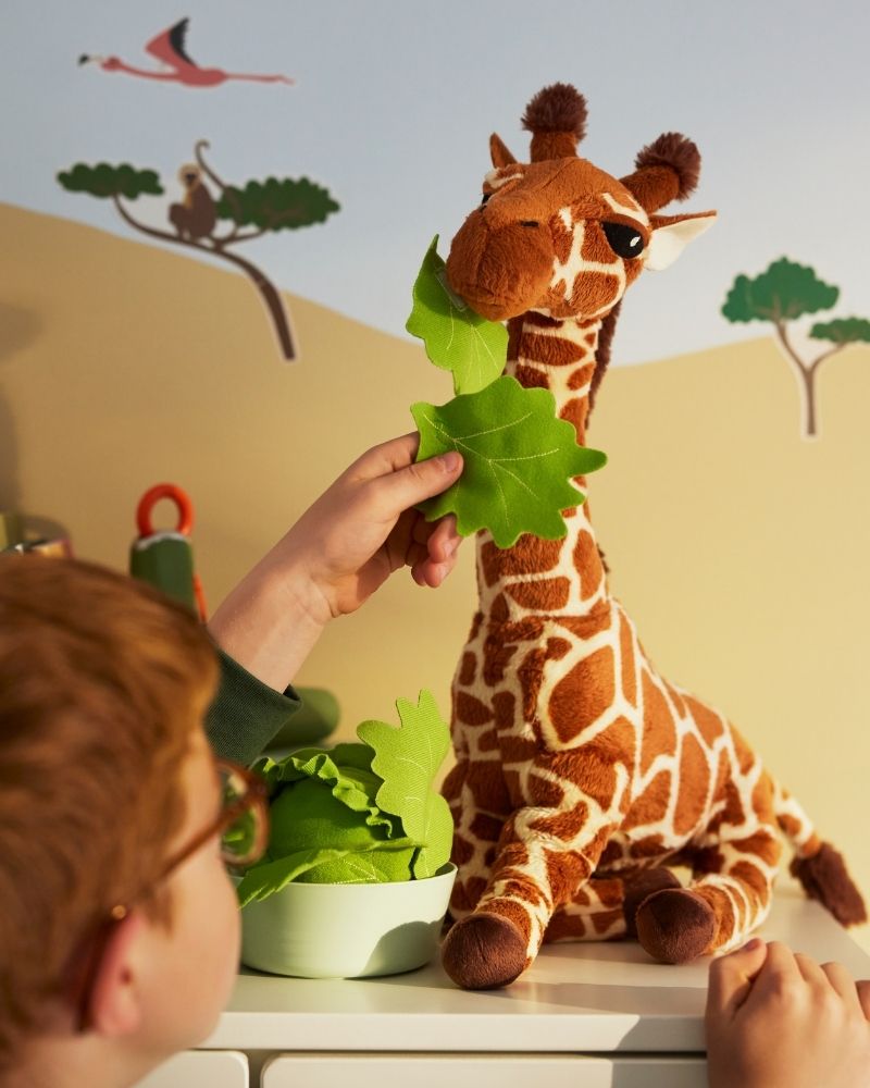 A child holds up a green leaf to feed a SANDLÖPARE soft toy giraffe.