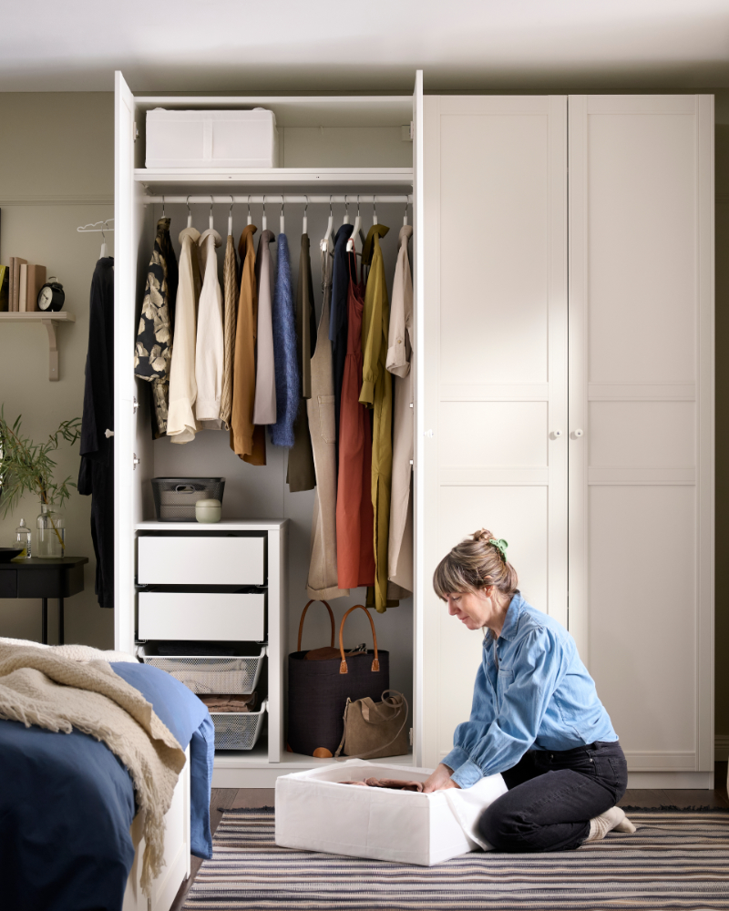Person organizing storage box in front of a white wardrobe with hanging clothes and drawers