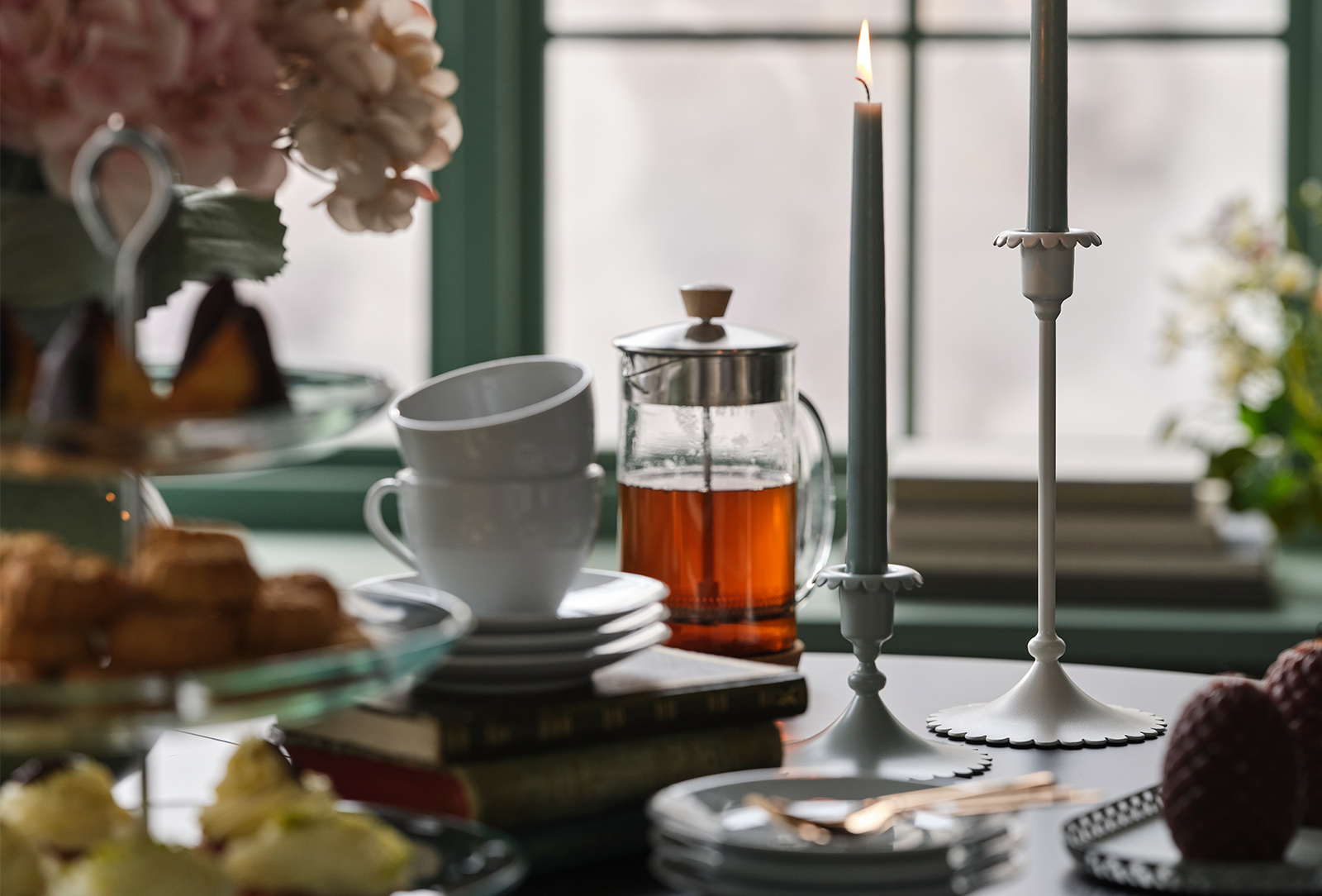 Two off-white SNÖSISKA candle holders stand on a dining table with tea and cakes in front of a green window.