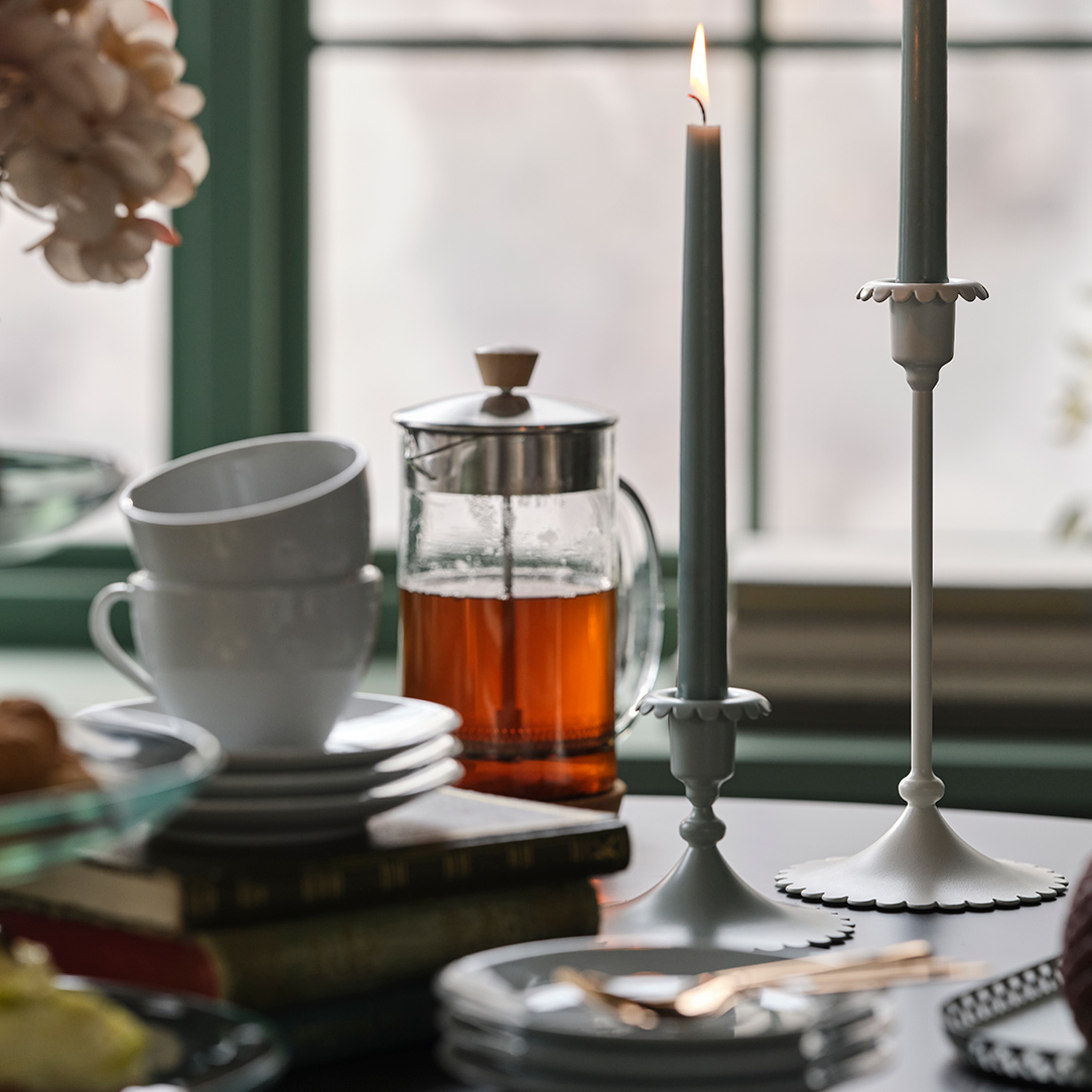 Two off-white SNÖSISKA candle holders stand on a dining table with tea and cakes in front of a green window.