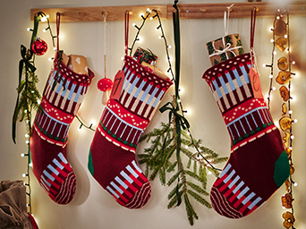 Tres calcetines navideños rojos y verdes colgados junto a ramas de abeto y luces decorativas.