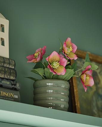 A FEJKA artificial potted plant inspired by a pink Christmas rose stands on a green shelf with books and paintings.