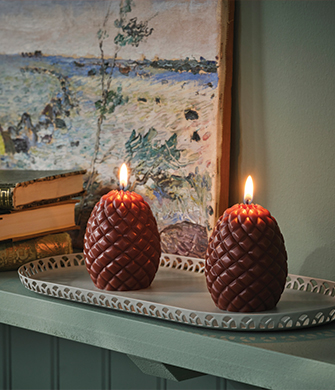 Dos velas encendidas en forma de piña de pino sobre una bandeja decorativa.
