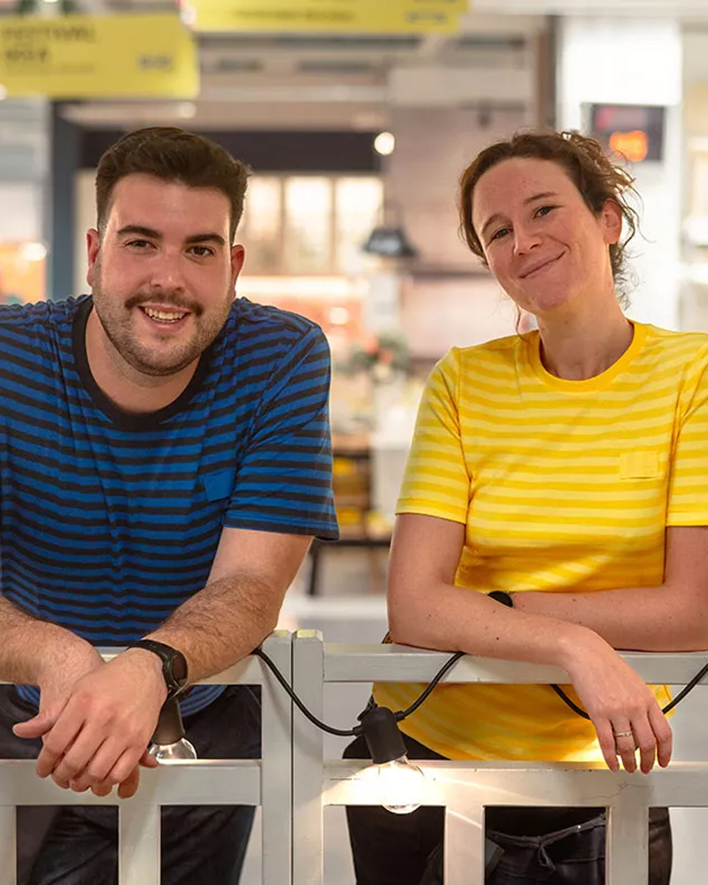 Two IKEA employees smiling in front of a white railing with string lights inside an IKEA store. The man wears a striped blue and black shirt, and the woman wears a yellow striped shirt.