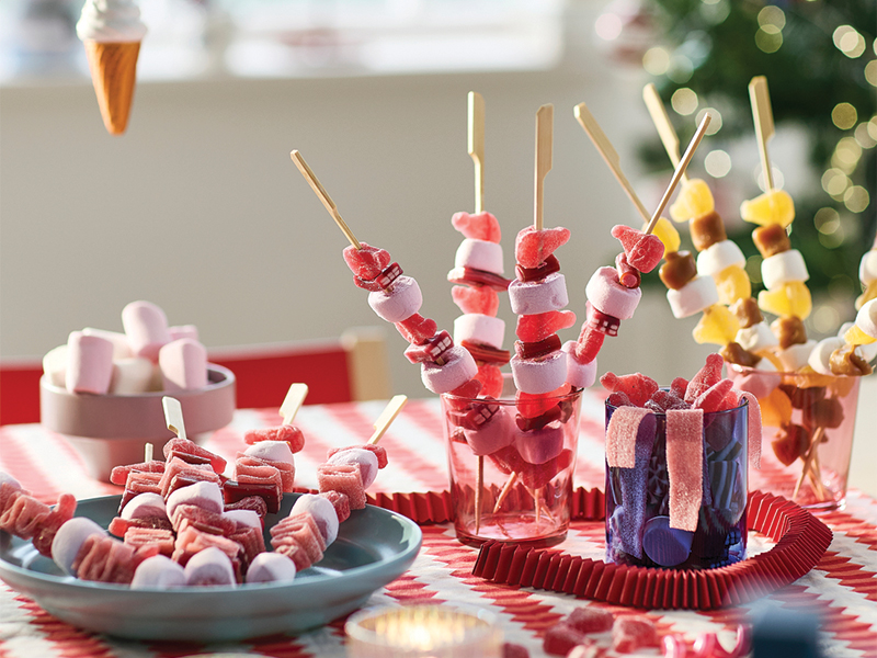 Candy and marshmallow skewers in glasses on a festively decorated table.