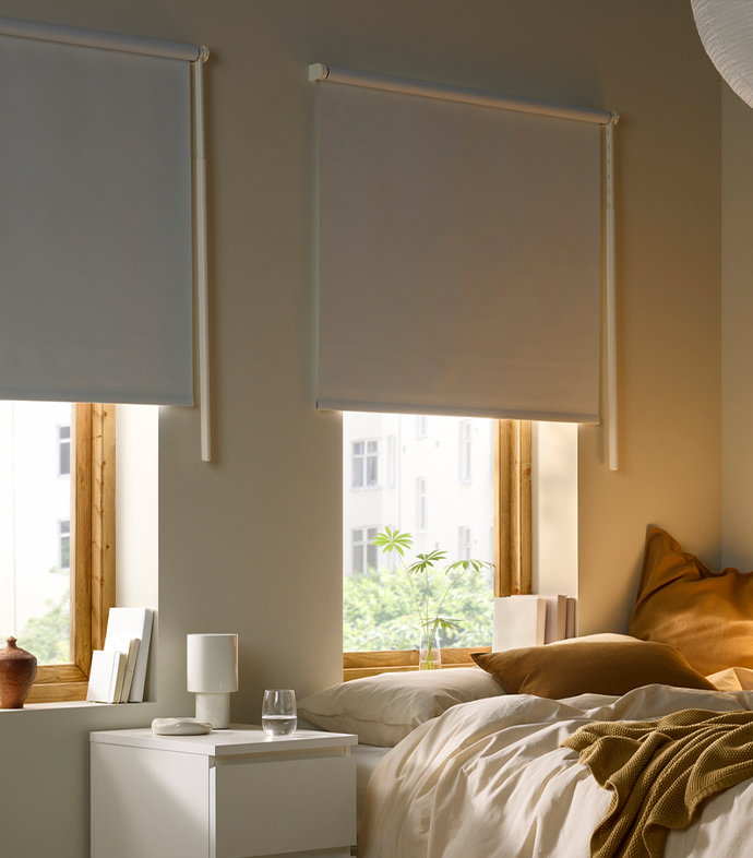 Cosy bedroom window area with white roller blinds pulled halfway down, a white bedside table, and a bed with warm ochre textiles.