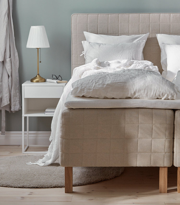 Cama tapizada en beige con ropa de cama blanca de lino junto a una mesilla blanca y una lámpara de latón con pantalla plisada.