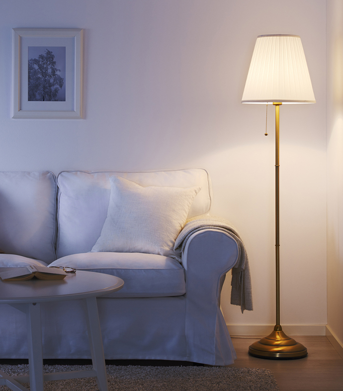 Rincón acogedor de salón con lámpara de pie en latón proyectando luz cálida junto a un sofá blanco con cojines neutros y mesa centro.